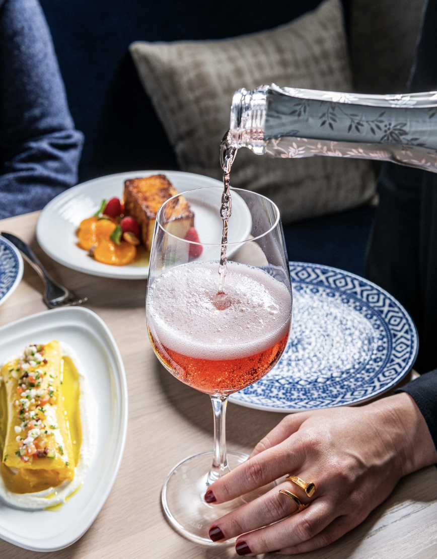 A hand holds a glass as sparkling rosé wine is poured, with plated desserts and a cushion in the background on a wooden table.