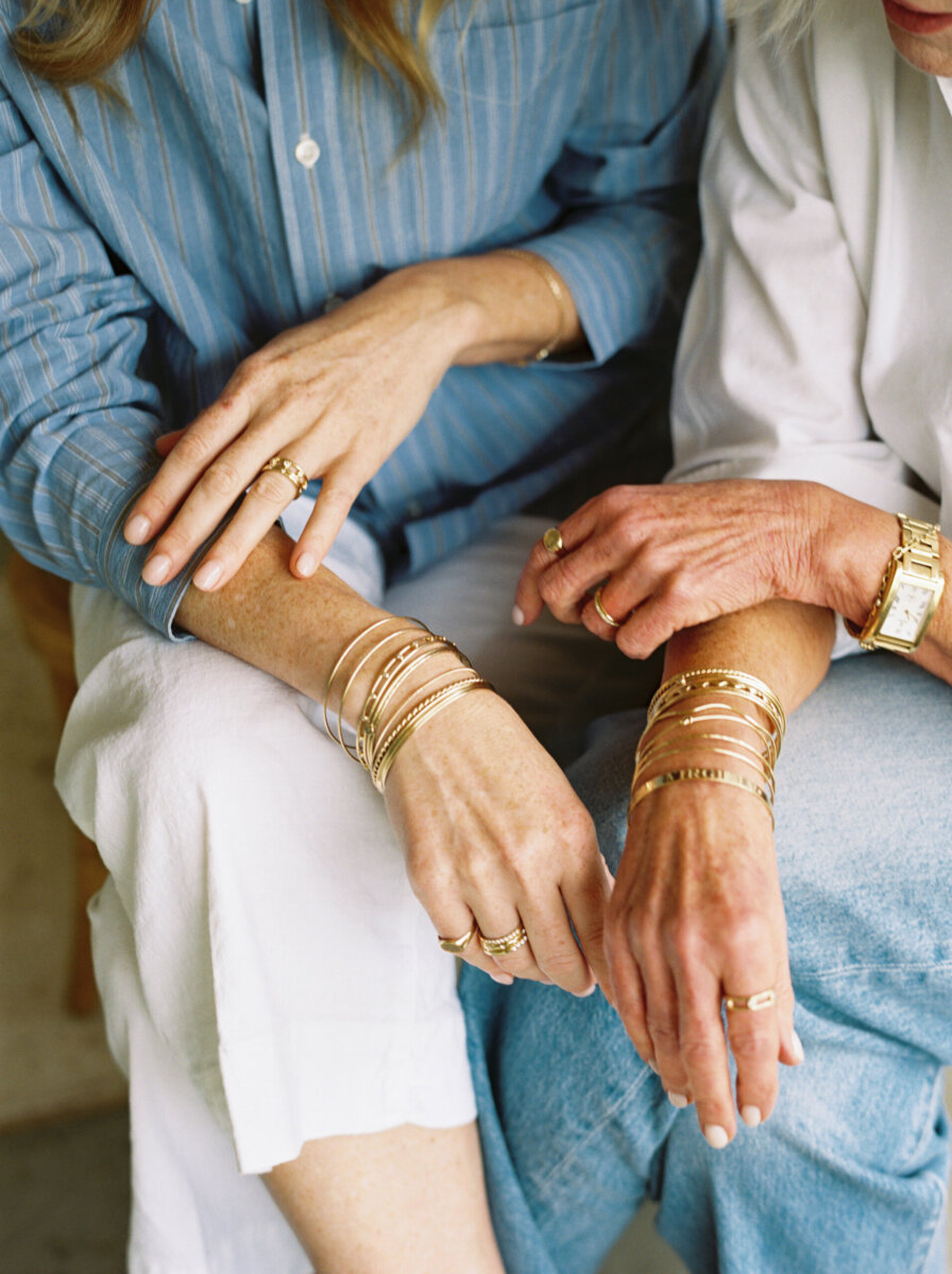 Two people sitting side by side, both wearing multiple gold bracelets and rings. They are dressed in light blue and white clothing, with their hands and arms prominently displayed—perfectly capturing a moment of style and elegance as the season’s fall savings arrive.