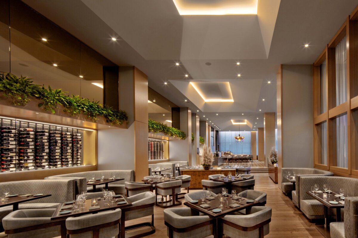 Elegant restaurant interior with modern lighting, wooden floors, gray upholstered seating, wine racks on the wall, and green plants as decor. Tables are set with glassware and cutlery.