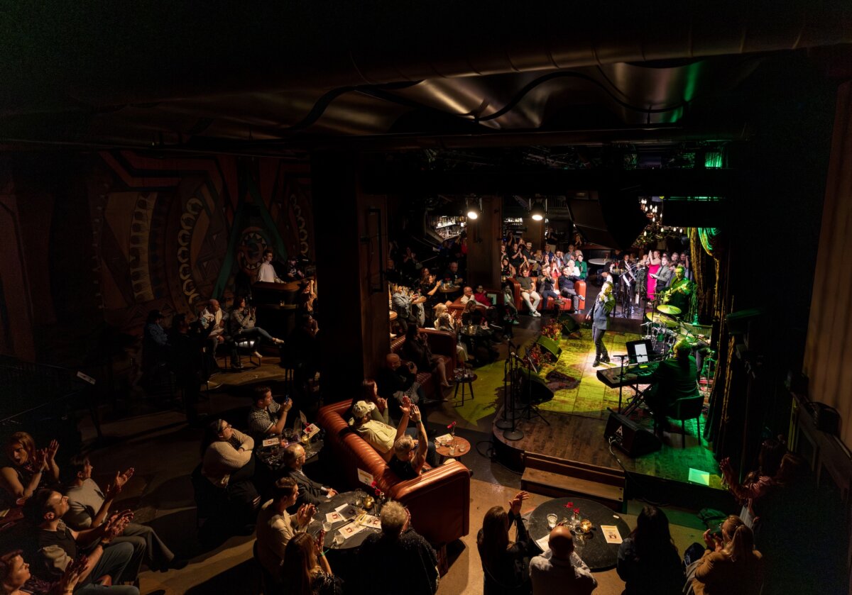 A band performs on stage in a dimly lit music venue while an audience sits at tables and applauds.
