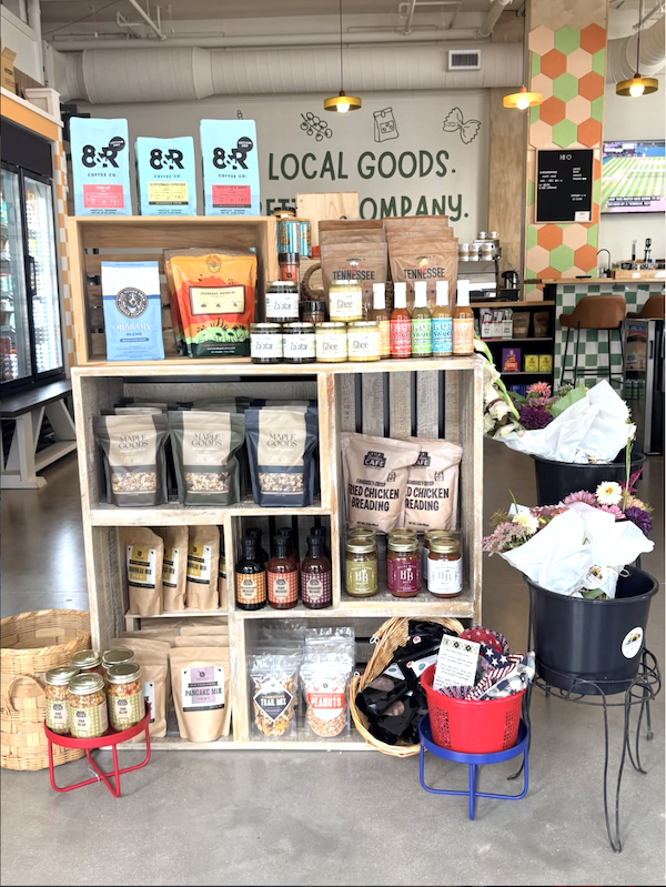 9 New Nashville Retail Spots to Explore - 5 Wooden shelves display assorted local food products, sauces, and snacks in a storeβan inviting example of Nashville shopping. Nearby, metal buckets hold flower bouquets. Wall text reads βLocal Goods. Etna Company.β.