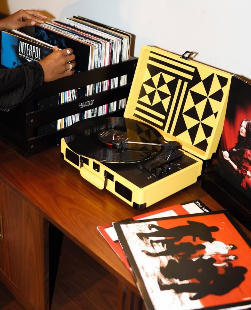 A hand selects a vinyl record from a crate next to a yellow record player on a wooden table in one of the cozy Nashville Vinyl Lounges, with album covers and a TV screen also visible—a quintessential 2026 music scene.