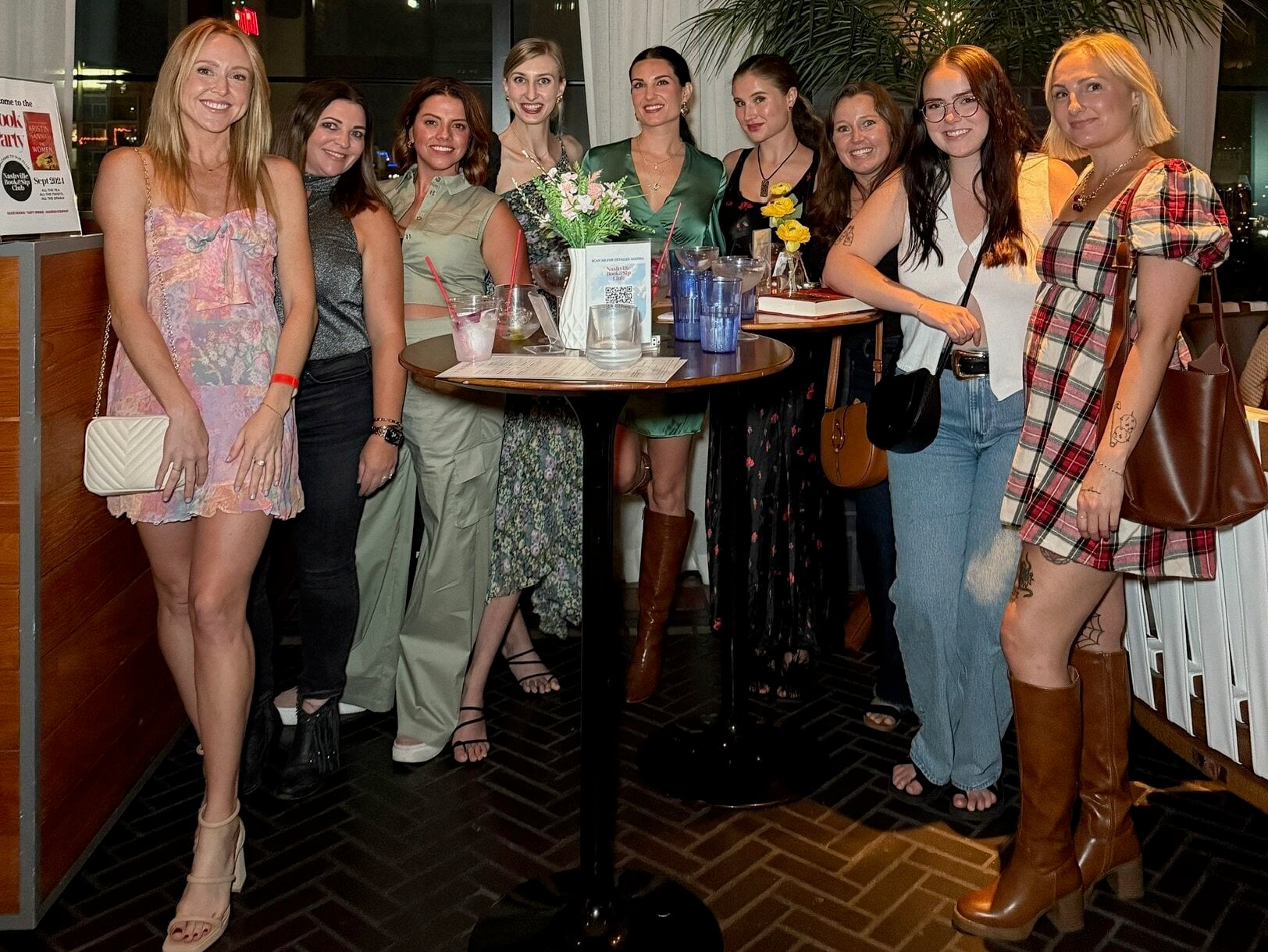 Nine women, members of one of Nashville’s vibrant women clubs, stand around a tall table with drinks and flowers, posing for a group photo in a modern indoor setting with large windows and lush plants.