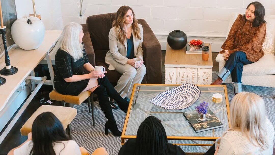 Six women sit in a modern living room engaged in conversation, arranged in a circle around a coffee table with decorative items—a warm gathering.