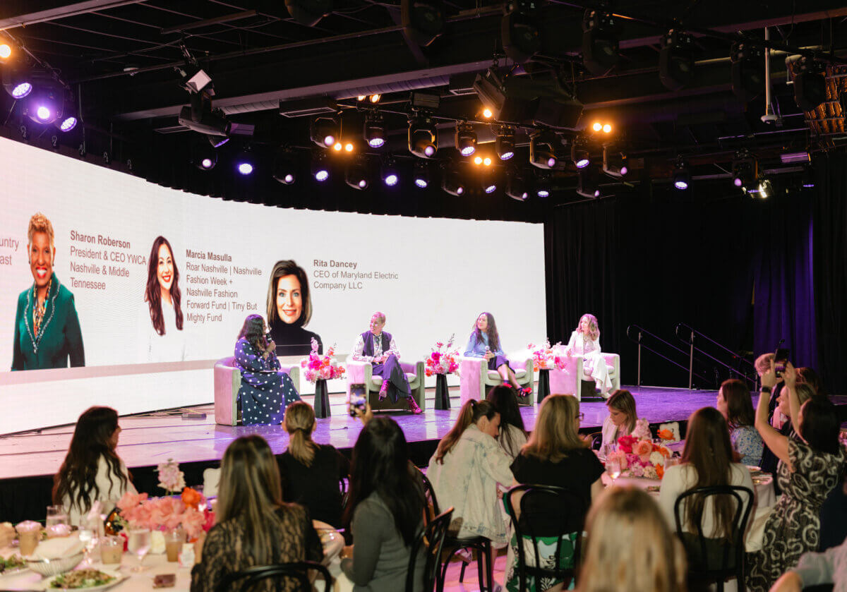 A panel of four women speaks on stage at a conference, representing some of the leading women’s clubs in Nashville, with their names and titles displayed on a large screen behind them. Audience members sit at round tables in the foreground, engaged in learning more about exciting Nashville women activities.