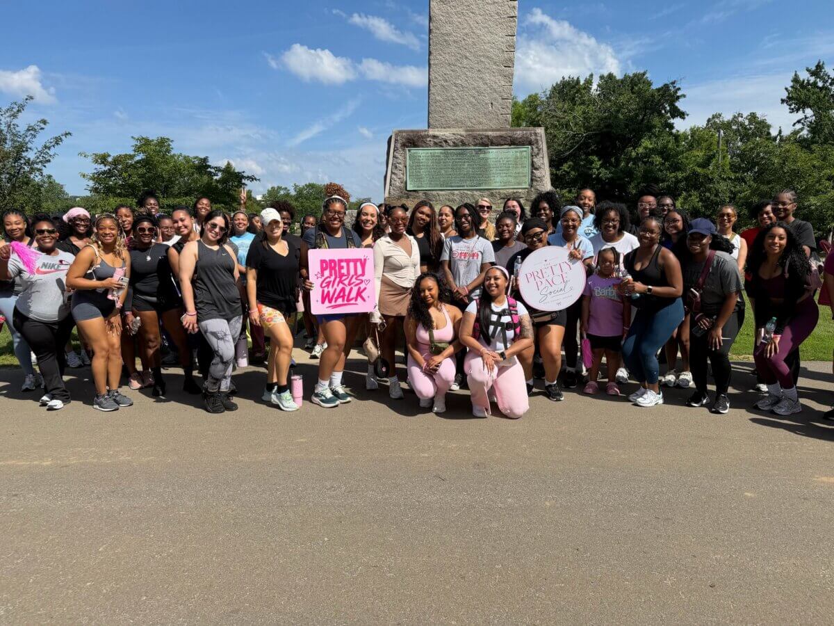 A large group of women poses together outdoors by a monument in Nashville, some holding signs that read “Pretty Girls Walk.” The weather is sunny and the group, gathered as part of special interest clubs, is dressed in athletic wear.