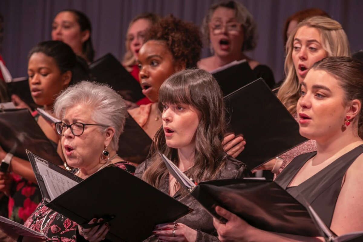 A group of women sing in a choir, each holding open black folders with sheet music, standing close together on stage.