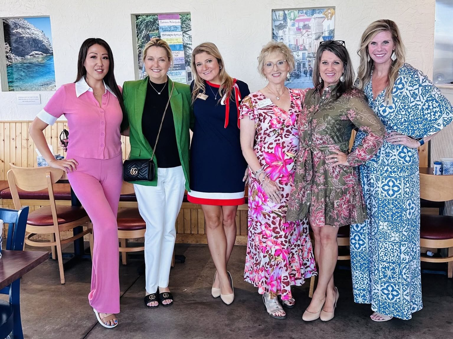 Six women stand indoors, posing and smiling in colorful outfits, with framed wall art and wooden chairs visible in the background.