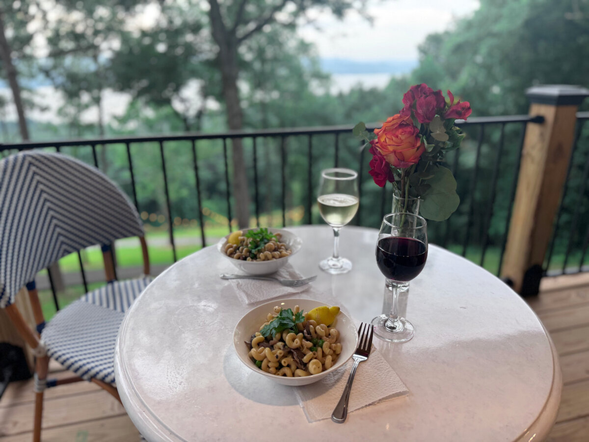Round table with two pasta bowls, a glass of white wine, a glass of red wine, and a vase of orange-red roses on an outdoor patio overlooking trees and water—an inviting scene perfect for enjoying lake views from your North Alabama glamping treehouse.