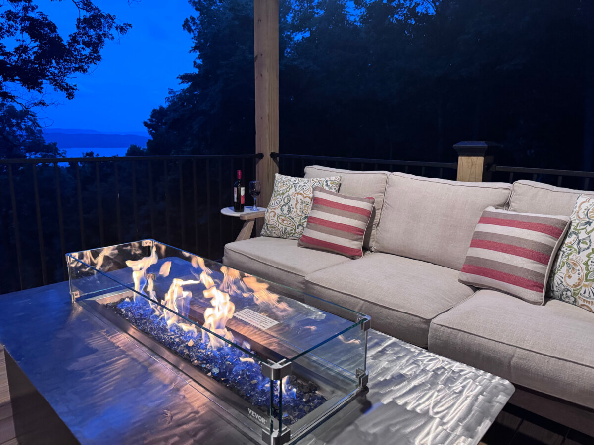 A modern outdoor seating area with a beige couch, striped pillows, a glass fire pit on a metal table, and a bottle of wine on a small side table at dusk—perfect for relaxing after a day exploring North Alabama glamping sites or taking in serene lake views from nearby treehouses.
