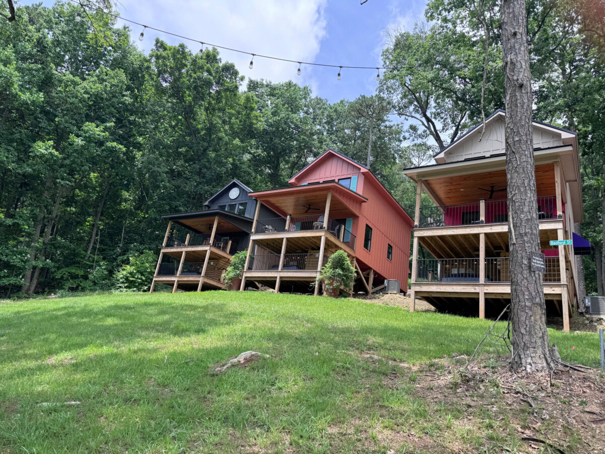 Three wooden cabins with covered porches stand side by side on a grassy slope, surrounded by trees under a partly cloudy sky—perfect for North Alabama glamping and soaking in peaceful lake views.