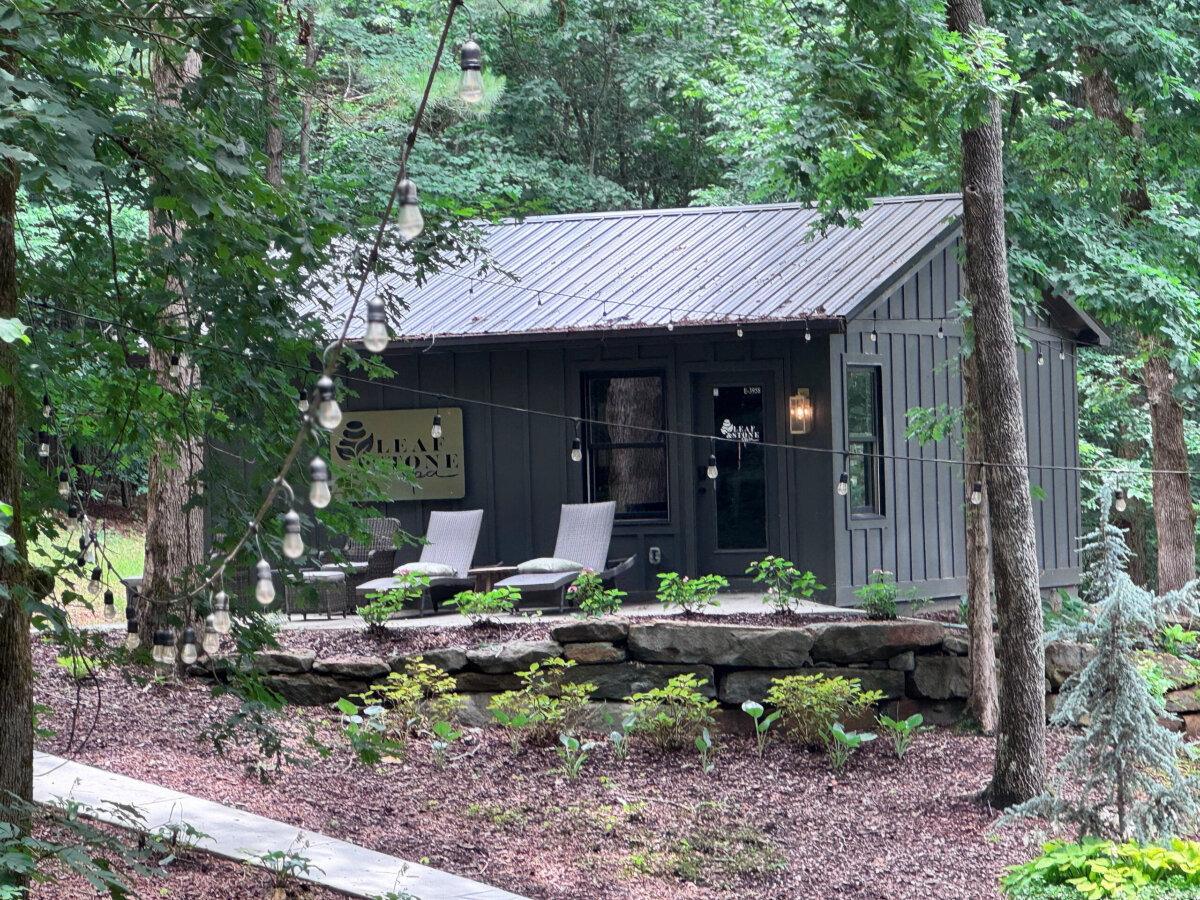 Small gray cabin with metal roof, surrounded by trees, has outdoor chairs on a stone patio and string lights overhead. Perfect for North Alabama glamping, this cozy retreat—named “Leaf Stone”—offers a peaceful forest setting just steps away from serene lake views.