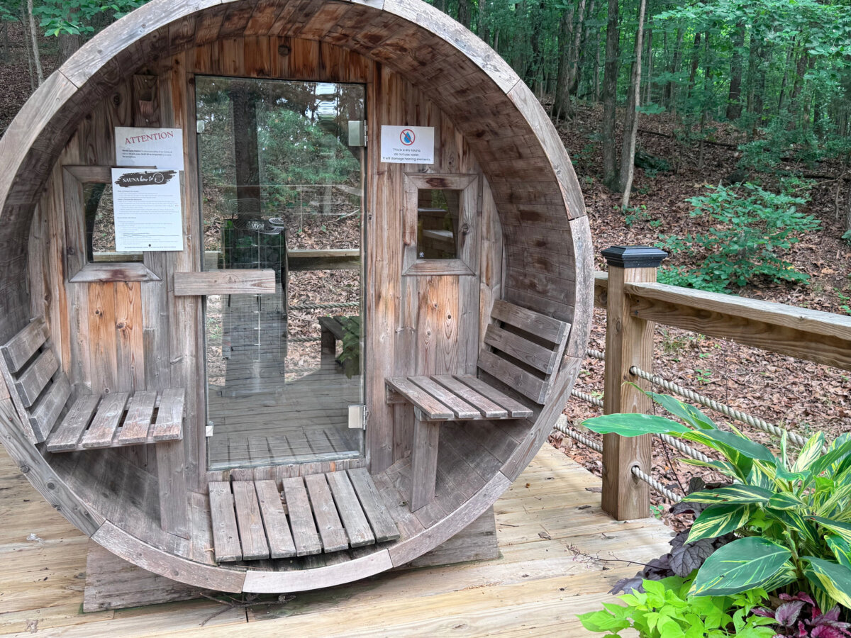 A round wooden sauna sits on a deck in a forested area, perfect for those seeking North Alabama glamping. Signs are posted on the door, and benches line the interior walls. Lush green plants are nearby, enhancing the peaceful atmosphere nestled among treehouses and lake views.