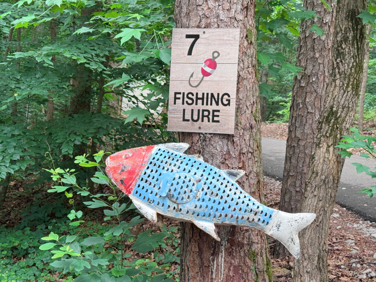 A wooden plaque with a fishing lure illustration and “7′ FISHING LURE” hangs on a tree above a large, painted fish sculpture in a wooded area—perfect for those seeking unique North Alabama glamping experiences surrounded by nature and lake views.