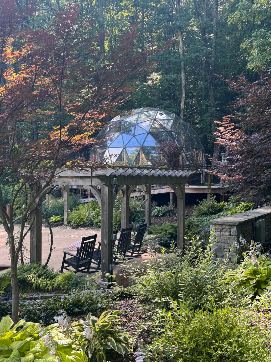 A glass geodesic dome sits behind a wooden pergola with rocking chairs, surrounded by lush greenery and trees in a garden setting, offering an enchanting North Alabama glamping experience.