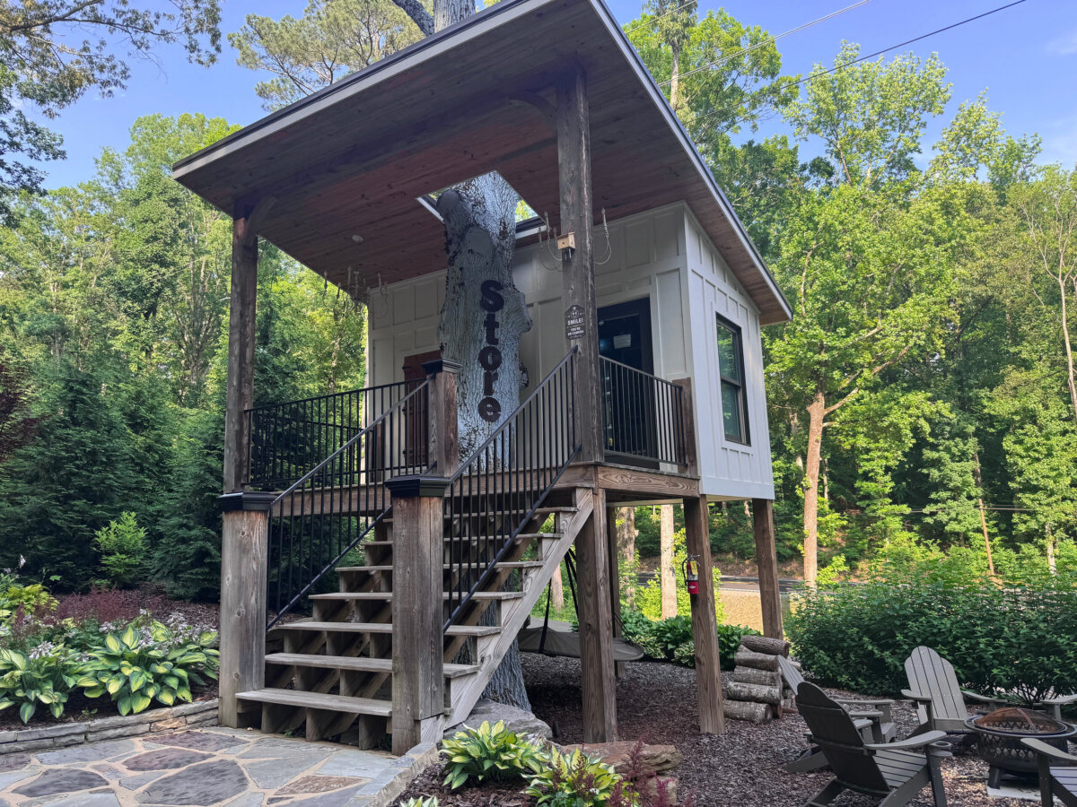 Elevated white cabin with wooden stairs, large porch, and “Store” sign, this inviting North Alabama glamping retreat is surrounded by trees, plants, outdoor seating, and offers charming treehouse vibes with glimpses of tranquil lake views.