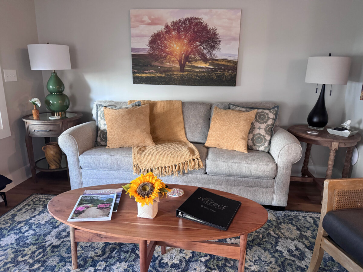 A living room with a gray sofa, yellow throw blanket, and pillows evokes the cozy charm of North Alabama glamping treehouses. A wooden coffee table holds magazines, a book, and a vase with a sunflower. Above the sofa hangs a tree artwork that hints at serene lake views just beyond your getaway retreat.