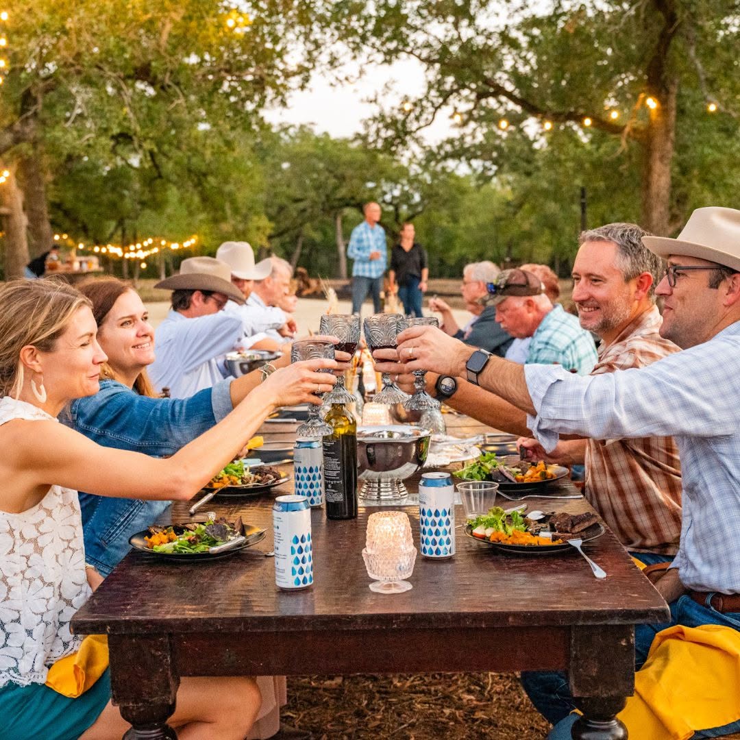 A group of people sit outdoors at a long table in the South, raising glasses in a toast, with food, candles, and string lights in the background—a perfect scene for those wondering where to find authentic live fire cooking.