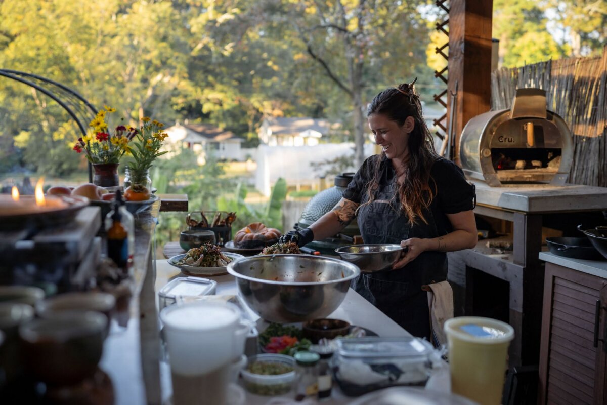 A woman prepares food at an outdoor kitchen counter surrounded by fresh ingredients, embracing the art of live fire cooking, with trees and houses in the background.