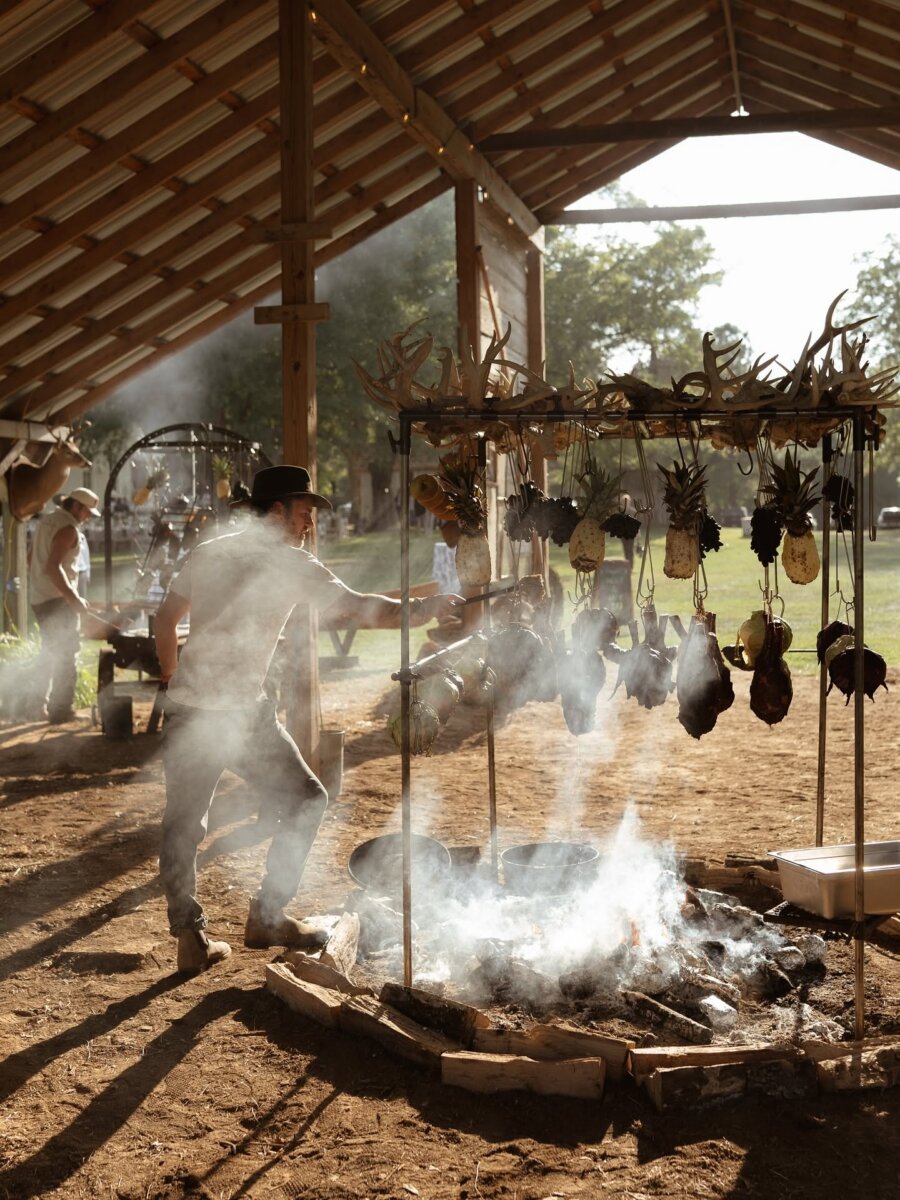 A person stands beside a smoky outdoor fire pit under a wooden roof, engaging in live fire cooking with meats and pineapples hanging above the flames. Another person works in the background, bringing the authentic flavors of Southern cuisine to life.