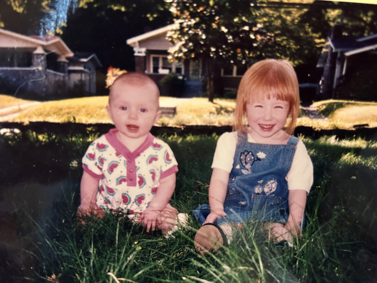 A baby and a young girl with red hair sit on grass in a sunlit Chattanooga yard, with houses and trees in the background—a charming scene capturing the creative force of childhood.