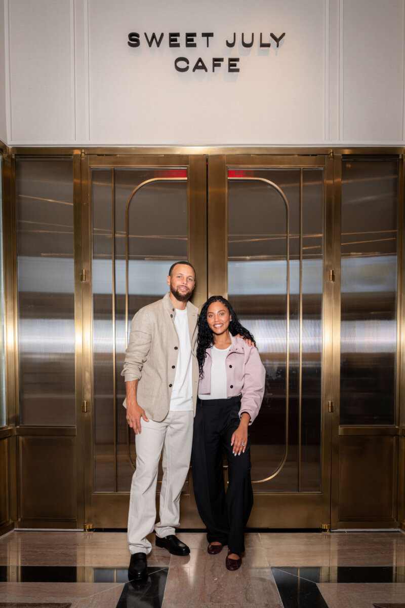 Two people stand smiling in front of the entrance to Sweet July Cafe, featuring gold doors and a marble floor.