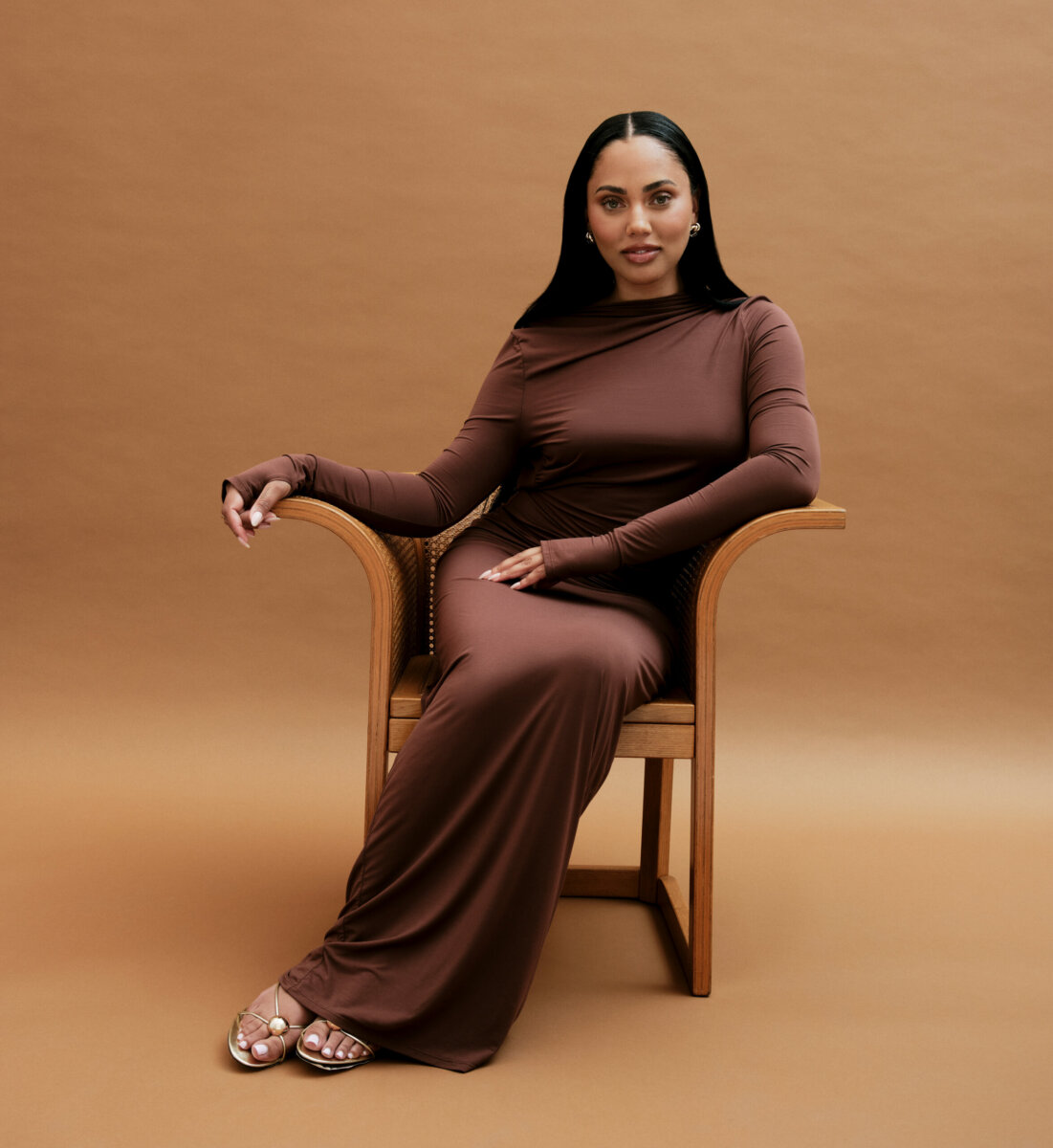 Ayesha Curry, with her long dark hair, sits in a wooden chair wearing a long-sleeved brown dress and gold sandals, set against an elegant brown backdrop.