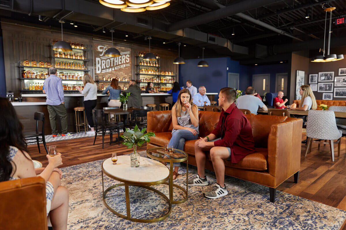 People sit and converse on leather couches in a modern bar with a stocked back wall and a sign reading “Brothers.” Several patrons stand at the bar, ready to raise a glass of Southern spirits; others are seated at tables around the room, making this one of the favorite stops for locals and visitors alike.