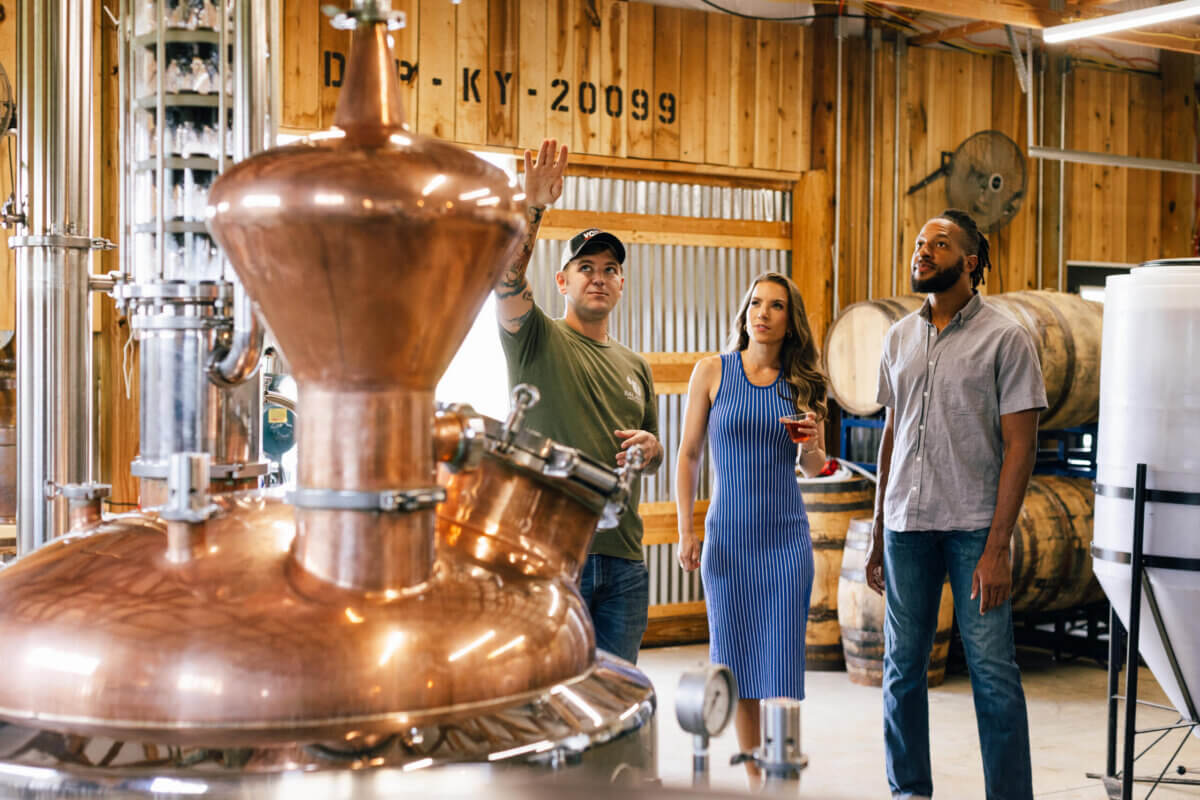 Raise a Glass: Our Favorite Stops for Southern Spirits - 13 Three people stand inside a distillery, listening to a man explain equipment near large copper stills and wooden barrels, as the rich tradition of southern spirits comes to life around them.