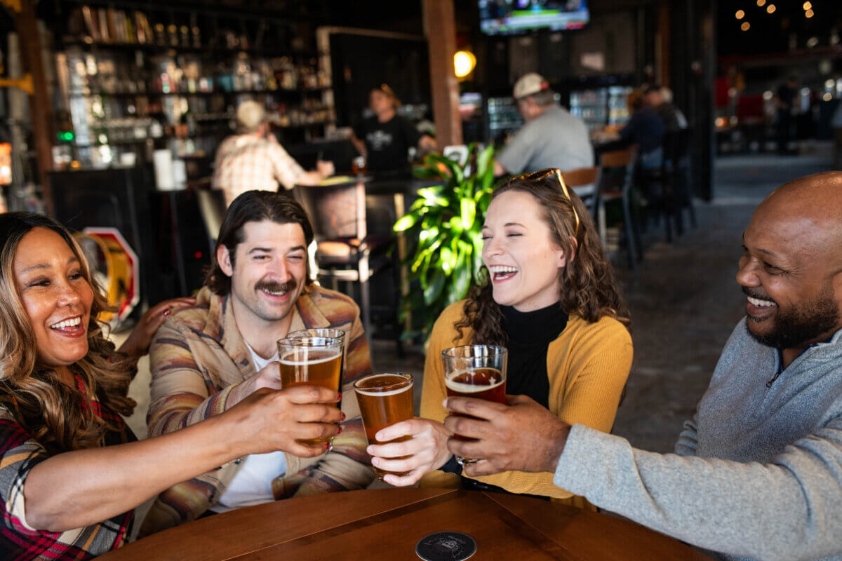 Raise a Glass: Our Favorite Stops for Southern Spirits - 12 Four people sitting around a round table clink glasses of beer and smile in a bar setting, with shelves lined with southern spirits and other patrons in the background.