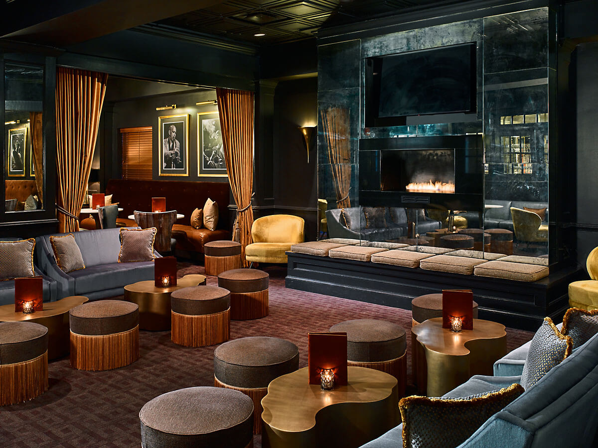 A modern lounge with plush seating, low round tables, golden accents, a fireplace, curtained alcoves, and framed art on dark walls—perfect for savoring southern spirits and expertly crafted southern cocktails.