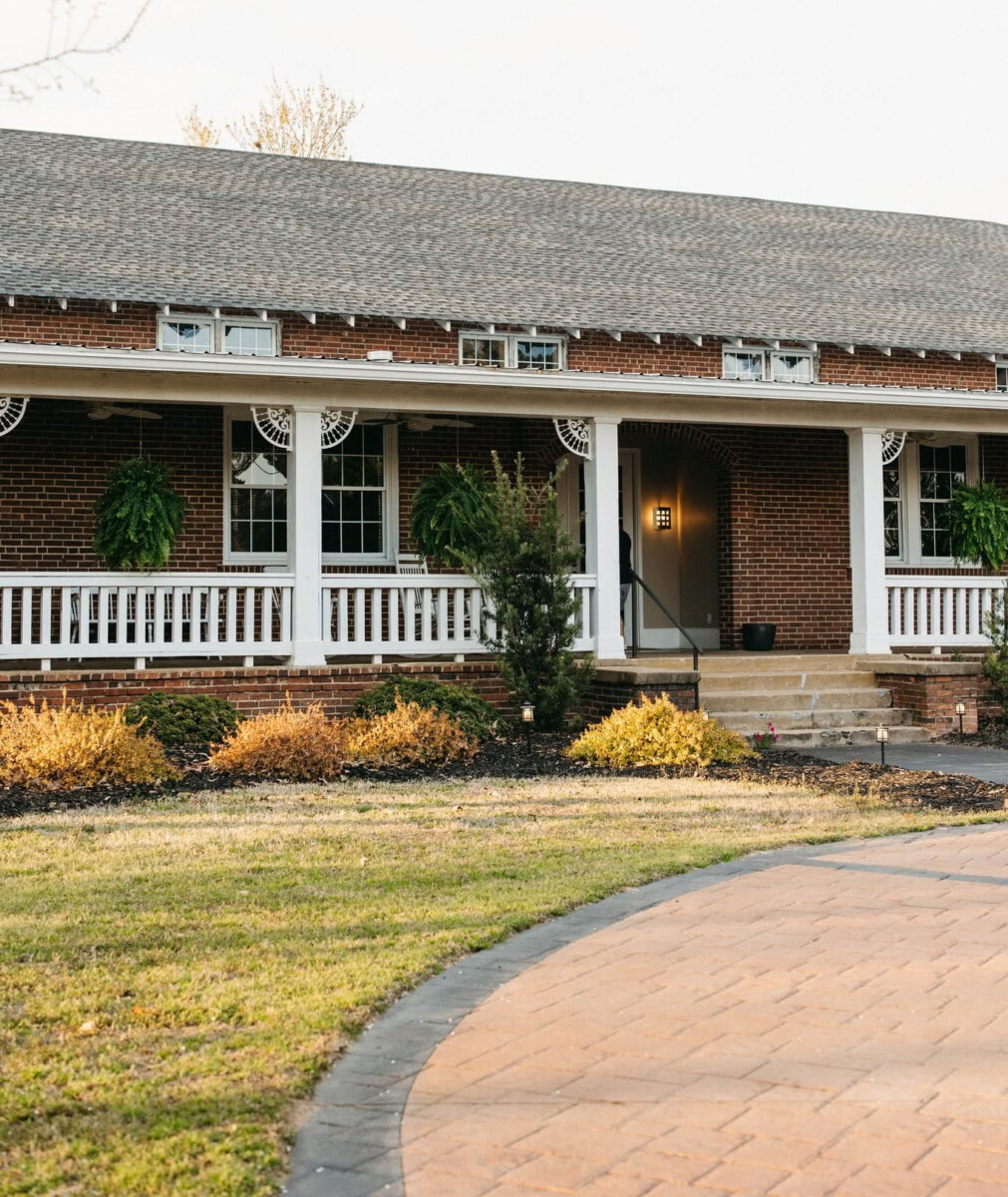 5 Destinations Where Southern Charm Thrives - 6 A brick house with a covered front porch exudes Southern charm, featuring white railings, hanging ferns, a black door, and steps leading to a curved brick walkway. The front yard is adorned with some shrubs and grass, creating an inviting scene that captures the essence of southern hospitality.