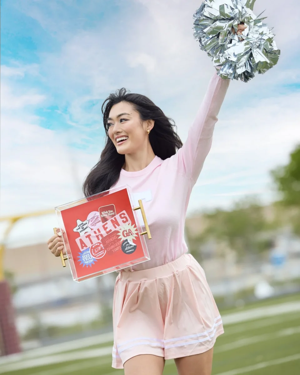 Southern Brands to Know for Football Season: Clothing, Decor & More - 9 A woman in pink athletic attire holds up a silver pom-pom and a red sign with βATHENSβ stickers, entertaining the crowd with her energetic spirit and colorful accessories while standing on a sports field.