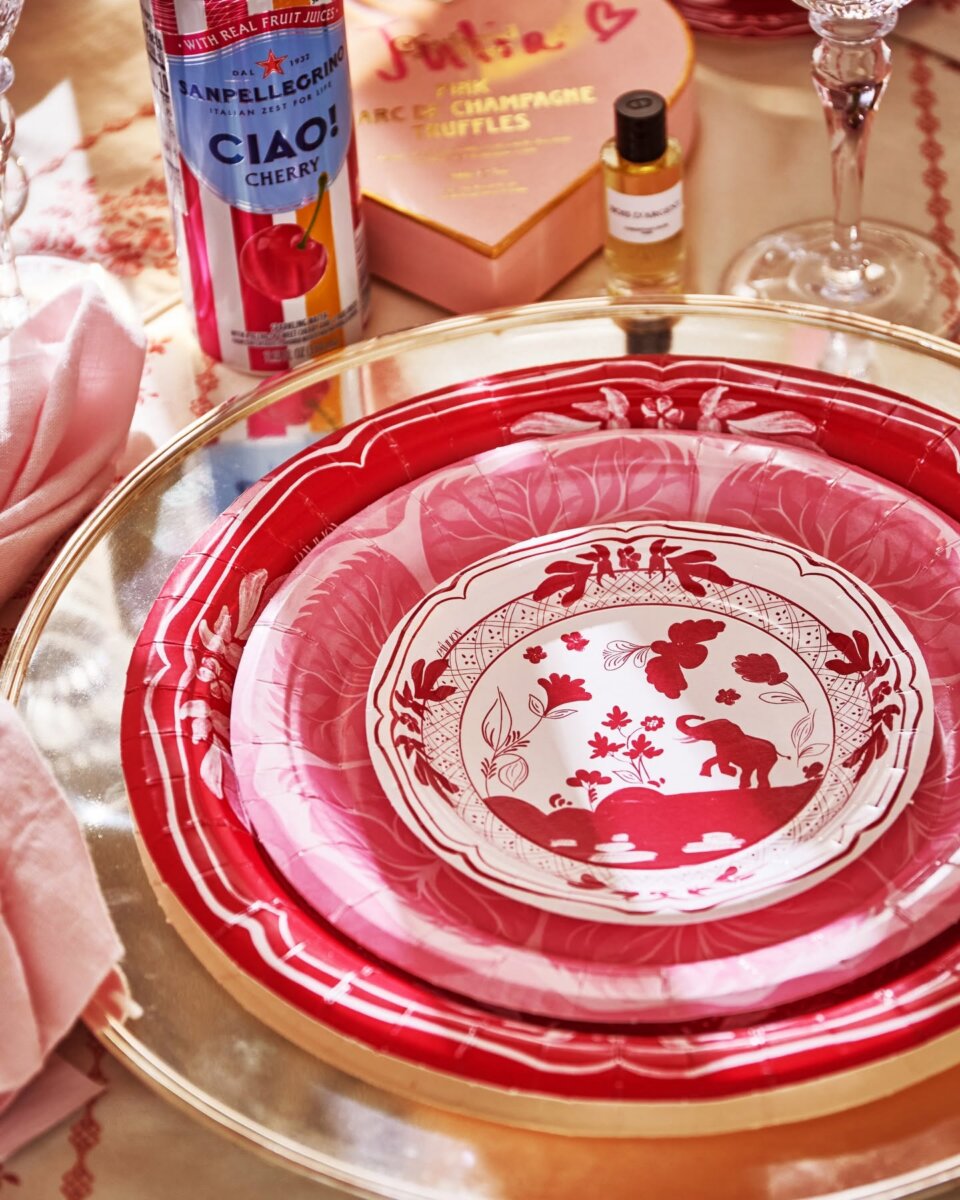 Southern Brands to Know for Football Season: Clothing, Decor & More - 8 A table setting with pink and red paper plates, a can of cherry Sanpellegrino, a box of champagne truffles, a small bottle, and pink napkins on a gold charger plateβperfect accessories for entertaining during football season.