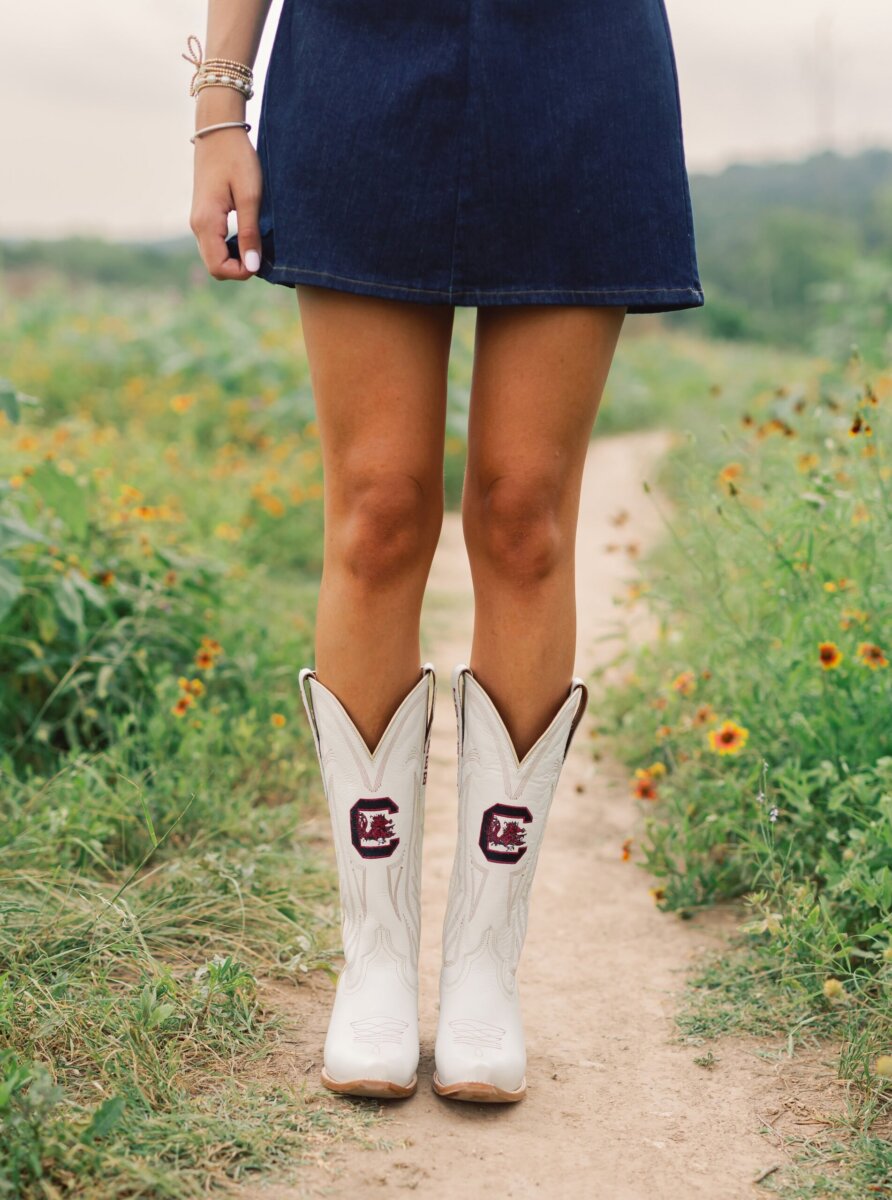 Southern Brands to Know for Football Season: Clothing, Decor & More - 6 Person standing on a dirt path wearing a denim skirt and white cowboy boots with embroidered maroon and black logosβperfect accessories for entertaining during football seasonβsurrounded by green grass and wildflowers.
