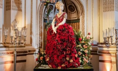 A mannequin in a ballroom at Cheekwood wears a gown made of red and white flowers, surrounded by elegant floral arrangements and golden decor.