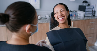 A woman smiles while sitting in a dental chair, wearing a protective bib, as one of the top dentists in Louisville, masked for safety, speaks to her.