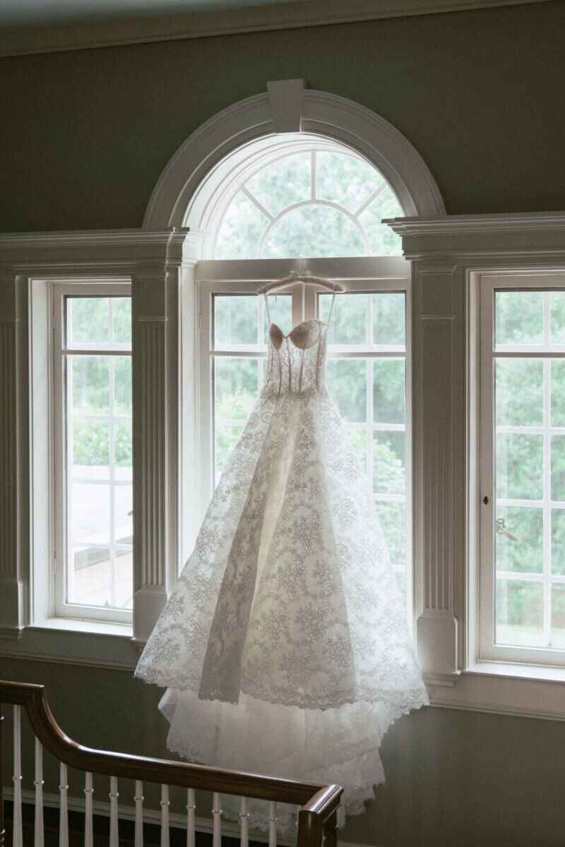 A Country Club Wedding with Show-Stopping Bridal Fashion & Peonies for Days - 4 A white lace wedding gown hangs in front of a large arched window at Belle Meade Country Club, with natural light streaming through and greenery visible outside—capturing the timeless elegance of a classic Nashville wedding.
