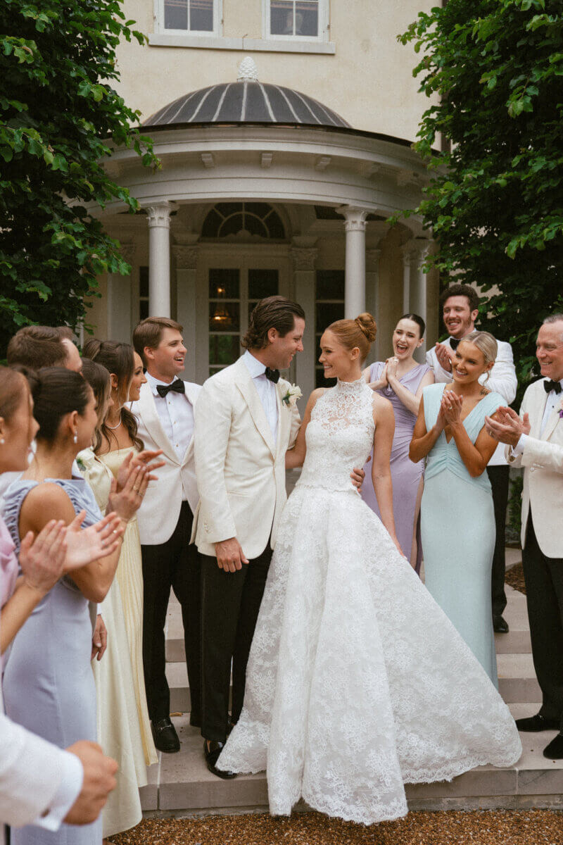 A Country Club Wedding with Show-Stopping Bridal Fashion & Peonies for Days - 2 A bride and groom stand together outside at Belle Meade Country Club, surrounded by their wedding party in formal attire, smiling and applauding as they celebrate a classic Nashville wedding.