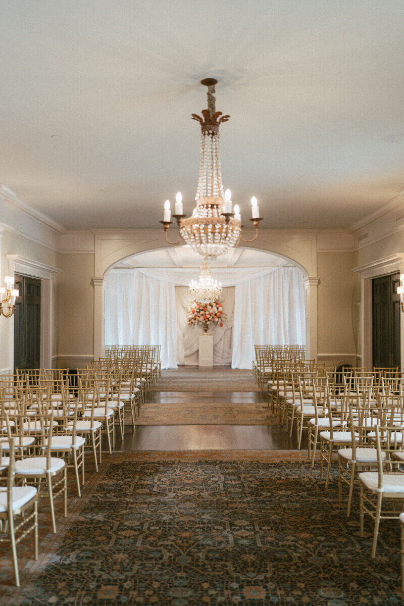 A Country Club Wedding with Show-Stopping Bridal Fashion & Peonies for Days - 16 A formal indoor wedding ceremony setup with rows of gold chairs, two chandeliers, white drapes, and a floral arrangement at the front creates an elegant atmosphere—perfect for a classic wedding at Belle Meade Country Club in Nashville.