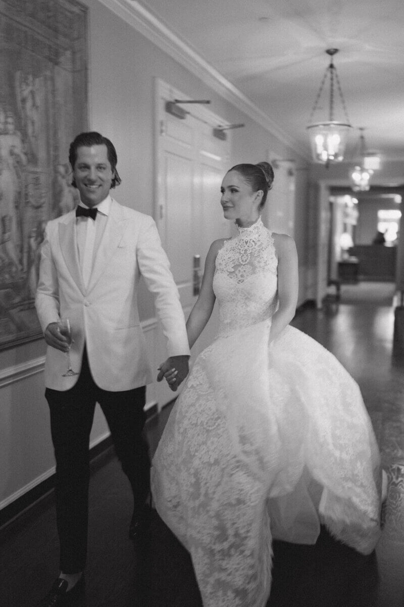 A Country Club Wedding with Show-Stopping Bridal Fashion & Peonies for Days - 3 A couple dressed in formal wedding attire walk hand in hand down a hallway at Belle Meade Country Club, the bride wearing a lace gown and the groom in a white tuxedo jacket, capturing the timeless elegance of a classic Nashville wedding.