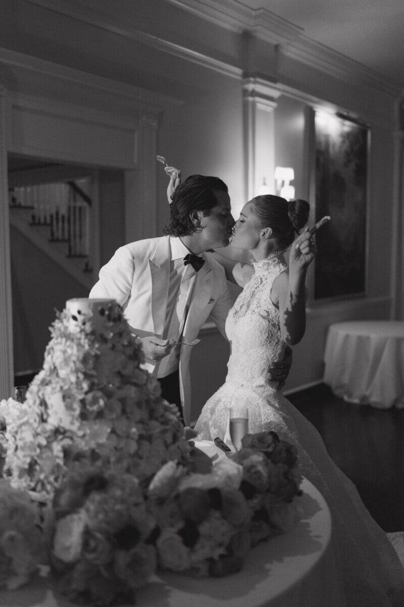 A Country Club Wedding with Show-Stopping Bridal Fashion & Peonies for Days - 23 A bride and groom in formal attire kiss while holding cake slices beside a large, flower-decorated wedding cake in an indoor venue, capturing the elegance of a classic wedding at Belle Meade Country Club.
