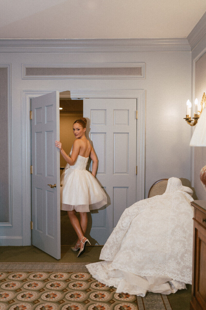 A Country Club Wedding with Show-Stopping Bridal Fashion & Peonies for Days - 6 A woman in a short white dress stands in a doorway, looking back and smiling. A long white gown is draped over a chair in a softly lit room with neutral walls and patterned carpet, capturing the timeless elegance of a classic wedding at Belle Meade Country Club.