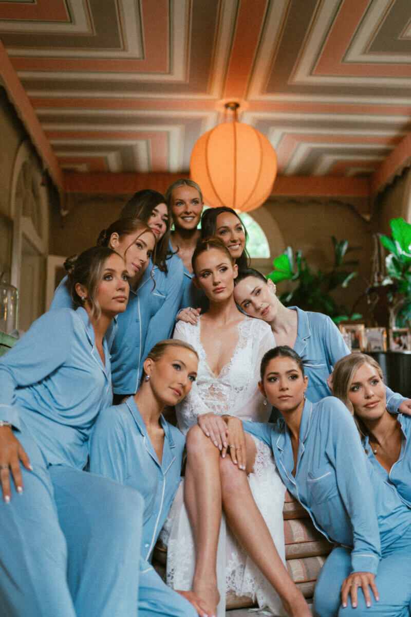 A Country Club Wedding with Show-Stopping Bridal Fashion & Peonies for Days - 9 A bride in a white lace dress sits surrounded by bridesmaids wearing matching light blue pajamas in a warmly lit room, capturing the timeless charm of a classic wedding at Belle Meade Country Club.