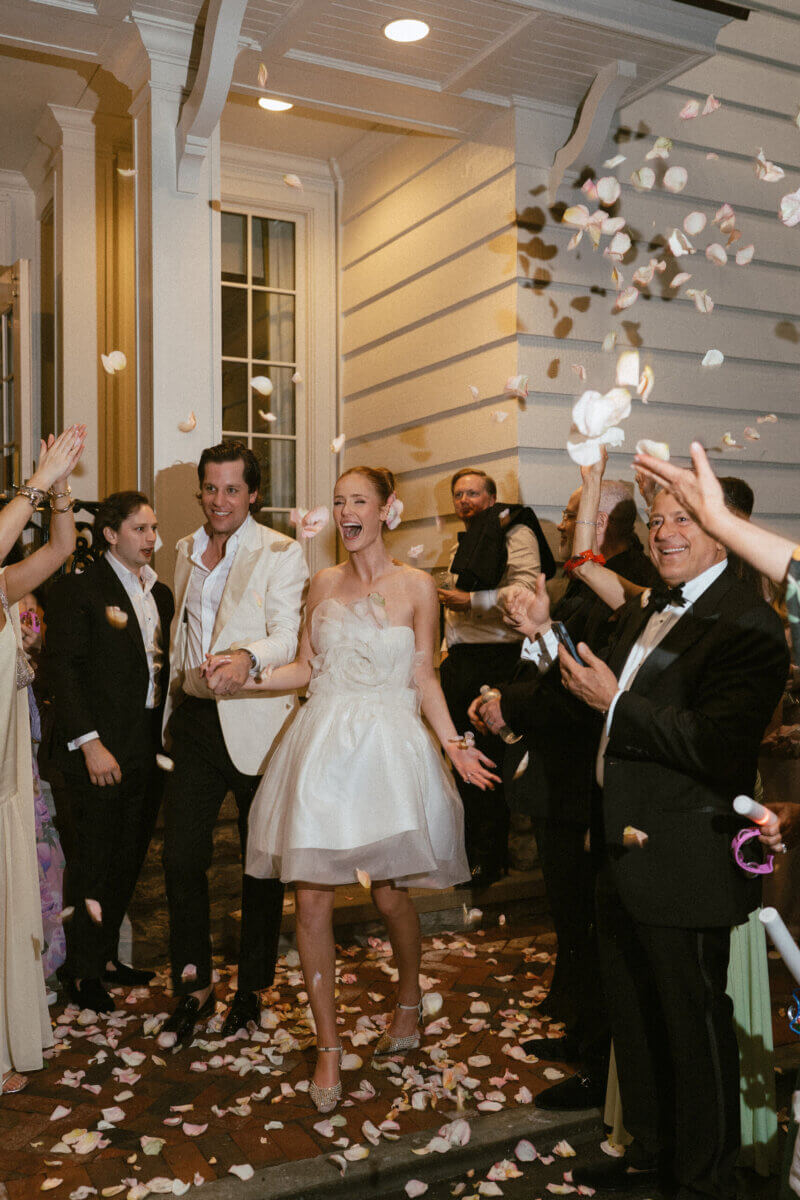 A Country Club Wedding with Show-Stopping Bridal Fashion & Peonies for Days - 26 A bride and groom walk outside at Belle Meade Country Club as guests throw flower petals and celebrate. The couple is smiling and dressed in classic wedding attire, capturing the joy of a gorgeous Nashville wedding.