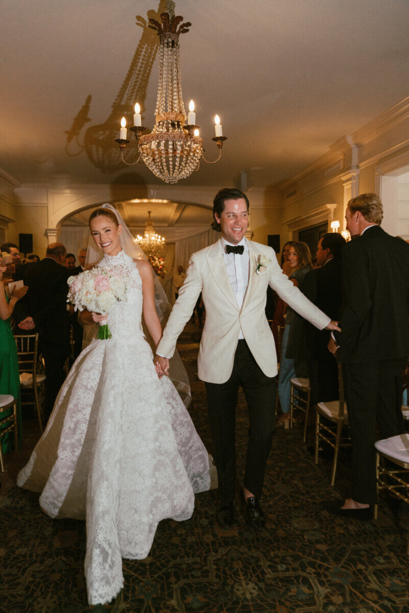 A Country Club Wedding with Show-Stopping Bridal Fashion & Peonies for Days - 17 A bride in a white gown and a groom in a white jacket walk down the aisle smiling and holding hands at an elegant indoor Nashville wedding, capturing the timeless charm of a classic wedding at Belle Meade Country Club.