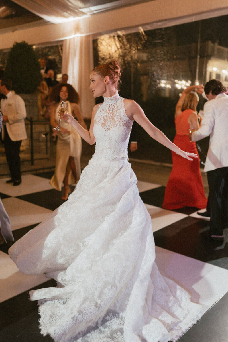 A Country Club Wedding with Show-Stopping Bridal Fashion & Peonies for Days - 24 At a classic wedding at Belle Meade Country Club, a woman in a white lace wedding dress dances joyfully on a black-and-white checkered floor, holding a champagne glass, while other guests join the celebration in the background.