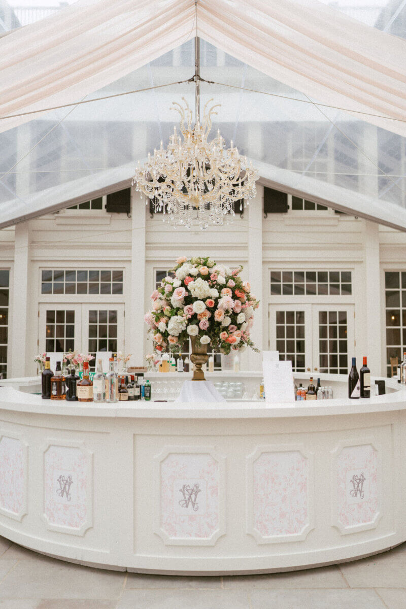 A Country Club Wedding with Show-Stopping Bridal Fashion & Peonies for Days - 12 White circular bar with assorted bottles, a large floral arrangement, and a crystal chandelier overhead under a clear tent roof at an outdoor venue—perfect for a classic wedding at the Belle Meade Country Club in Nashville.