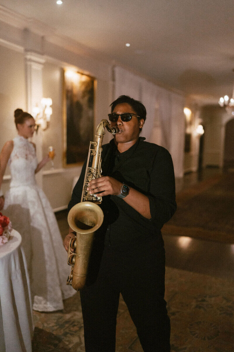 A Country Club Wedding with Show-Stopping Bridal Fashion & Peonies for Days - 25 At a classic wedding at the Belle Meade Country Club, a person wearing sunglasses and black clothing plays a saxophone indoors at the event; another person in a white dress stands in the background holding a drink.