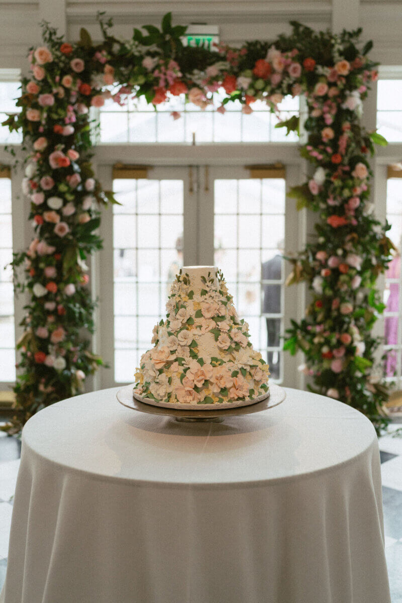 A Country Club Wedding with Show-Stopping Bridal Fashion & Peonies for Days - 22 A tiered white cake decorated with pink and green floral accents sits on a round table, framed by a large floral arch and glass doors in the background, creating an elegant setting perfect for a classic wedding at Belle Meade Country Club.